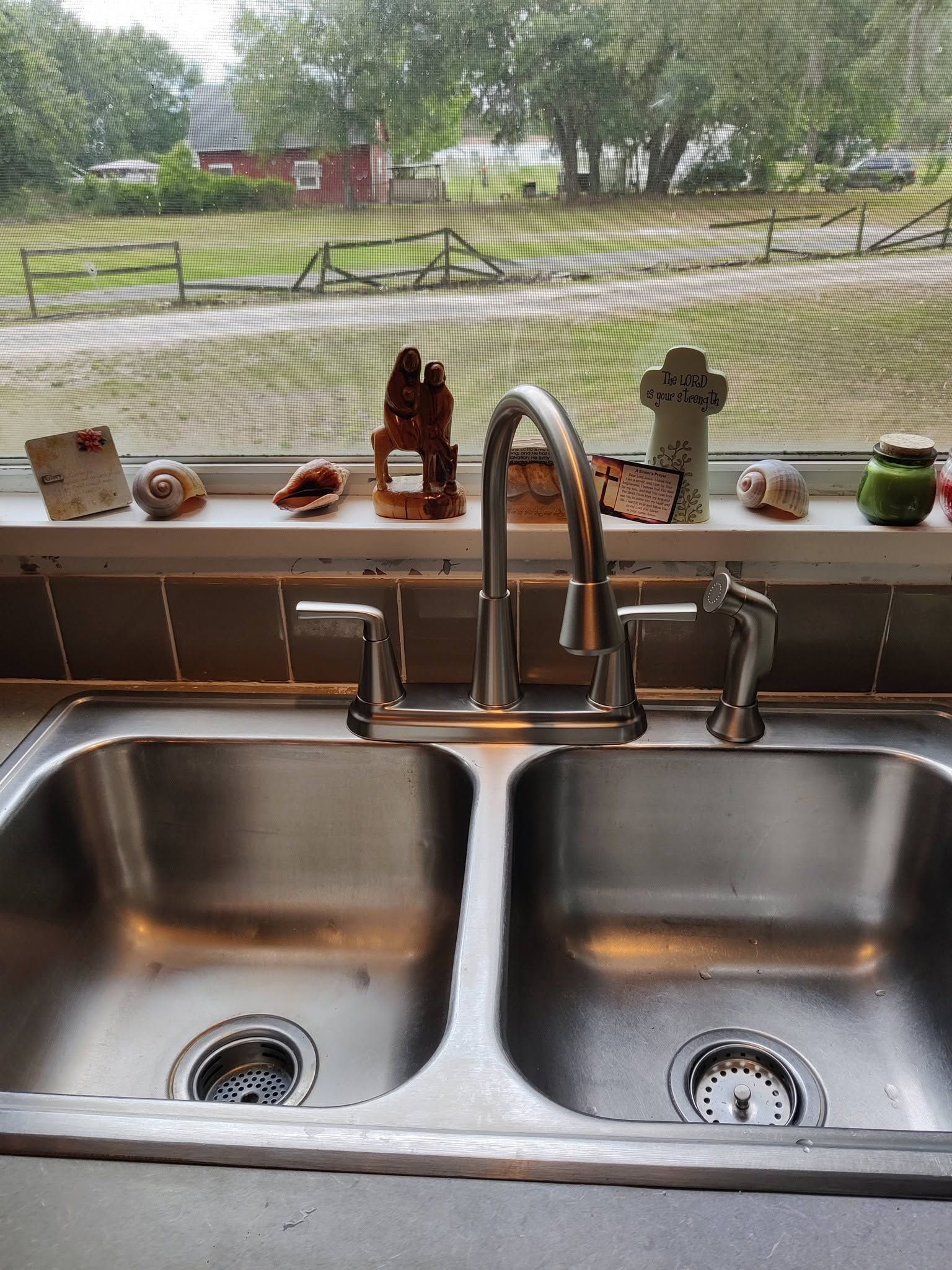 Kitchen sink with double basins and faucet, looking out to a rainy yard through a window.