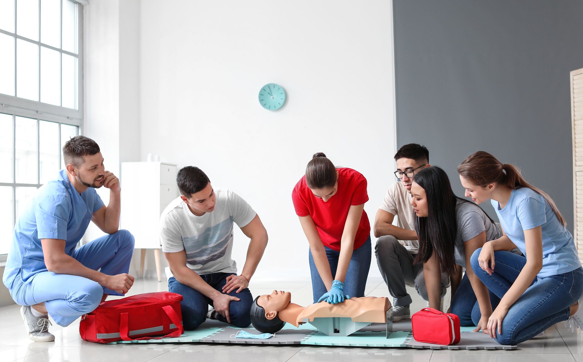 People practicing CPR on a training manikin in a bright room, with red first-aid bags nearby