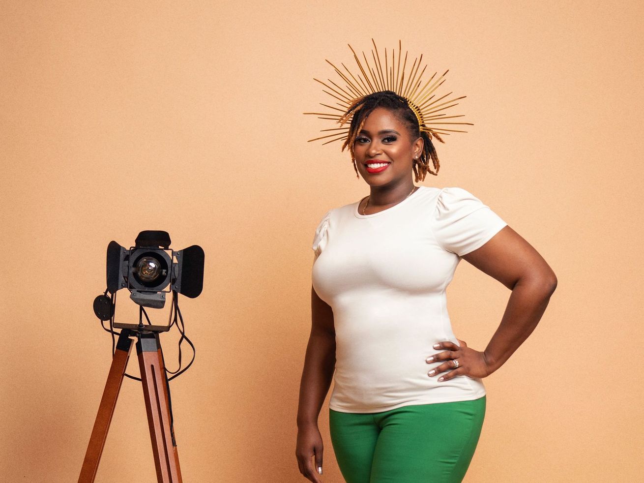 Woman posing in front of camera on tripod. Wearing white shirt, green pants, and a gold headpiece. Beige backdrop.