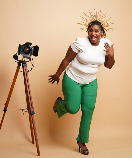 Woman posing next to a camera on a tripod, wearing a white shirt and green pants, gold headpiece.