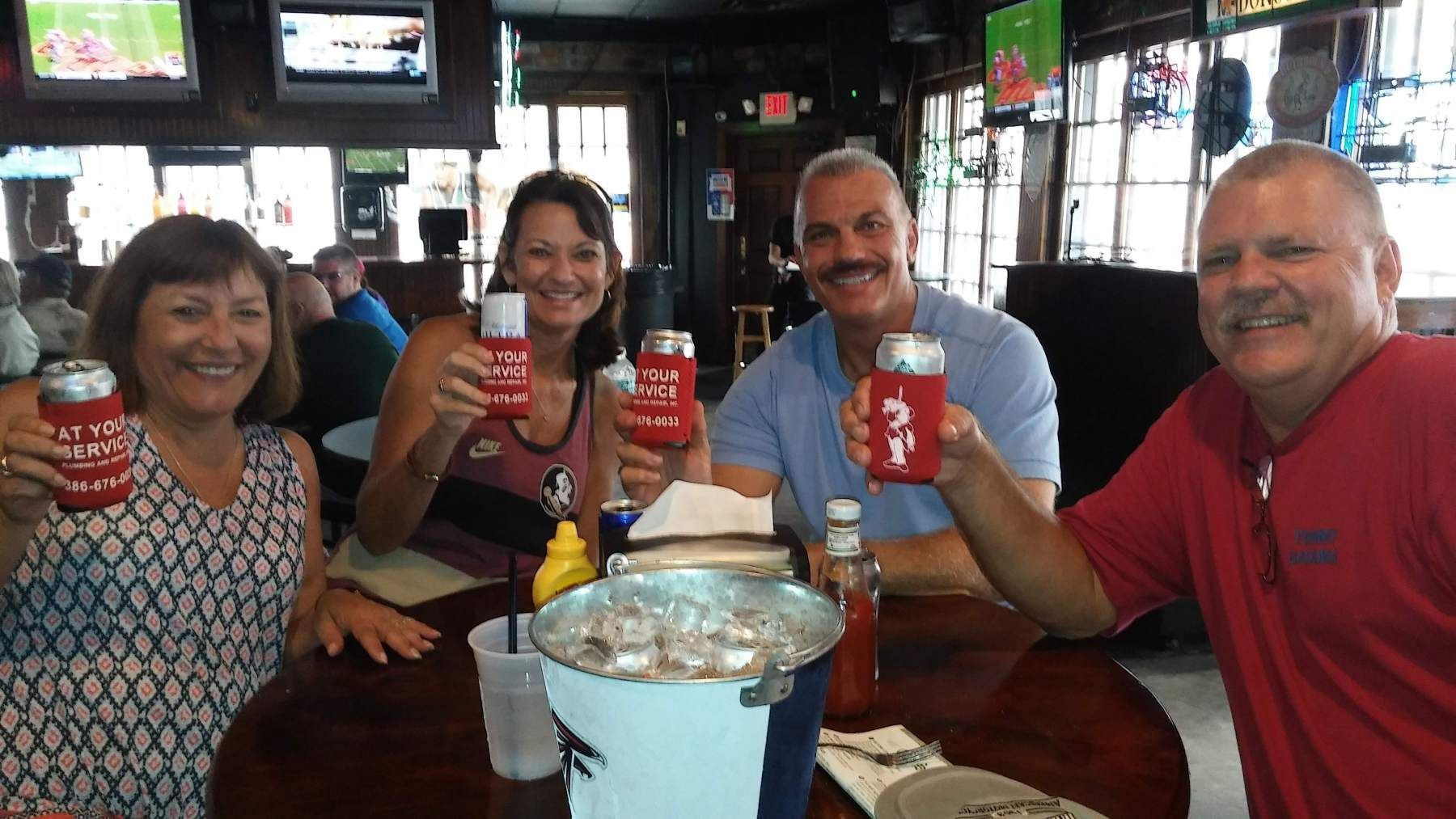 Four people at a bar cheer, holding up red drink containers. Bucket of oysters on the table.