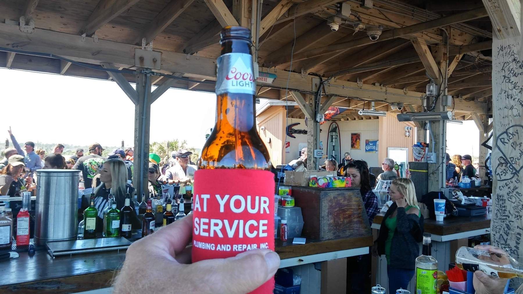 Person holding beer bottle with a red koozie at an outdoor bar with a crowd.