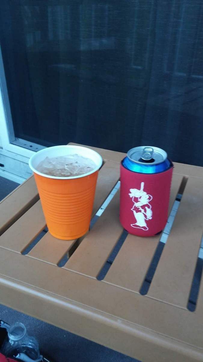 Orange cup of drink and red can in a koozie on a slatted brown surface.