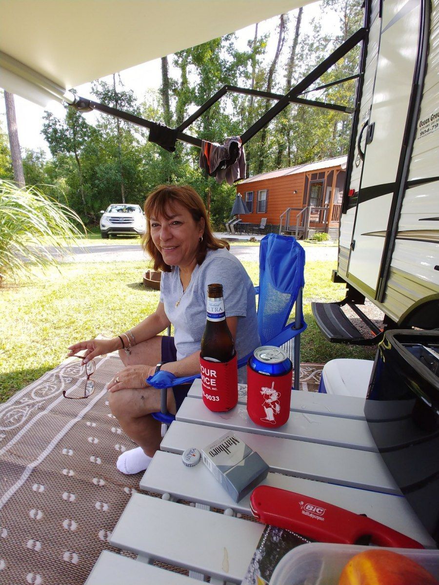 Woman sits at a table near a camper, holding a drink, outdoors.