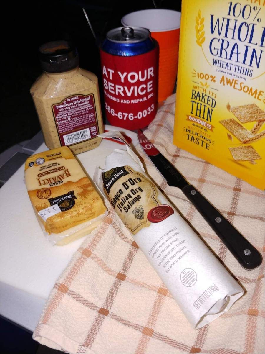Various food items: mustard, crackers, cheese, salami, and a drink, on a checkered cloth.