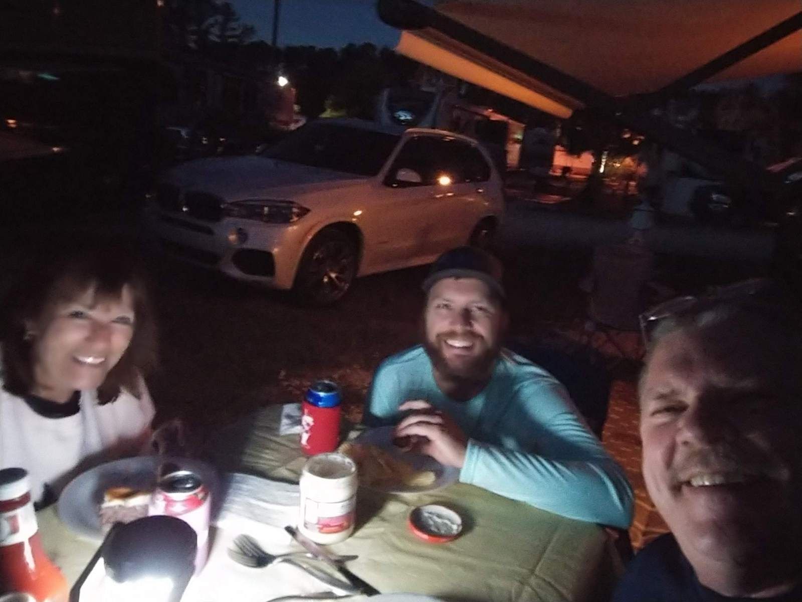 Three people smiling around a table at night with a white car in the background; camping or a cookout setting.