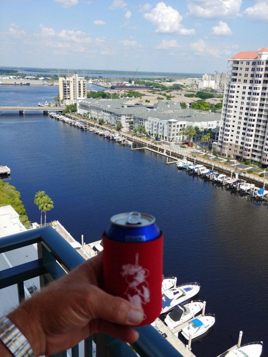 Hand holding a red can cooler with a city skyline and waterway in the background.