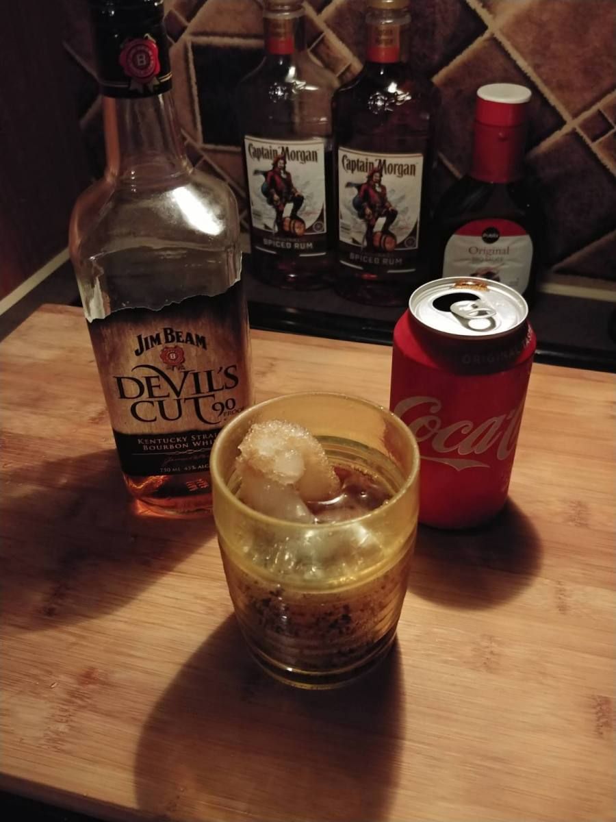 Cocktail with ice, Coca-Cola can, and rum bottles on a wooden surface.