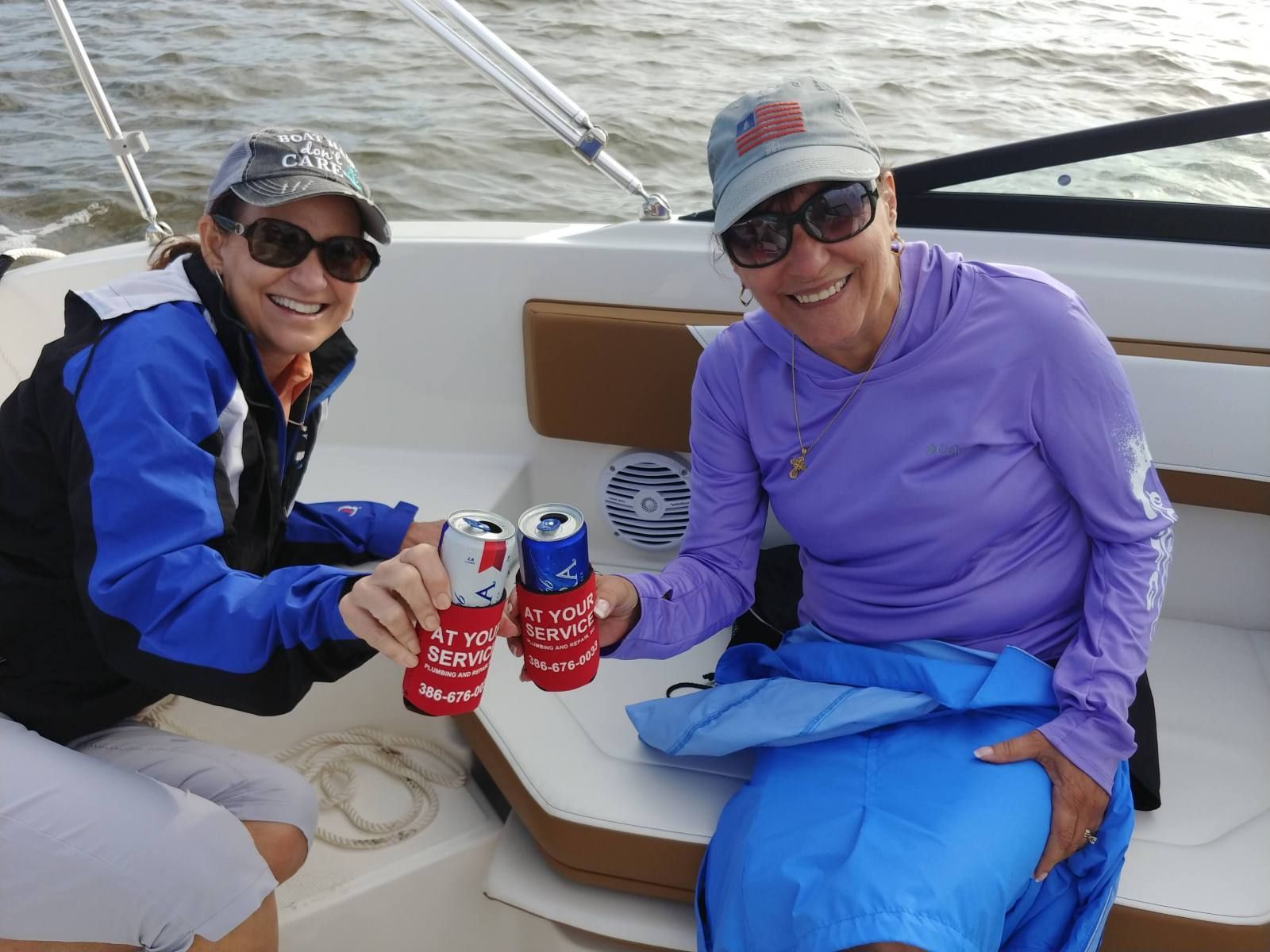 Two women on a boat raise cans of beer, smiling. One wears a blue jacket, the other a purple shirt. Ocean background.