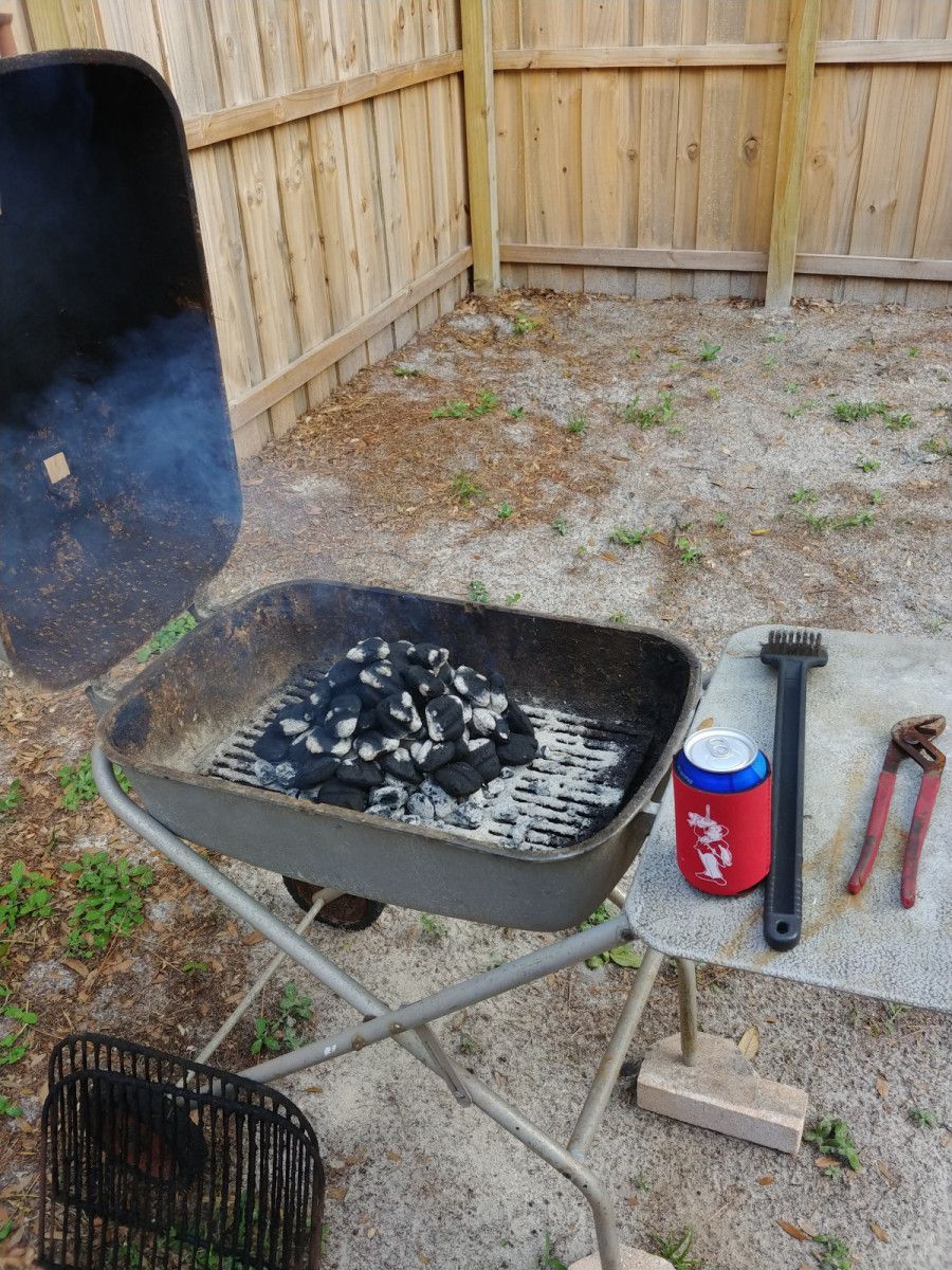 Charcoal grill with lit coals, lid open, next to a table with tools and a drink.
