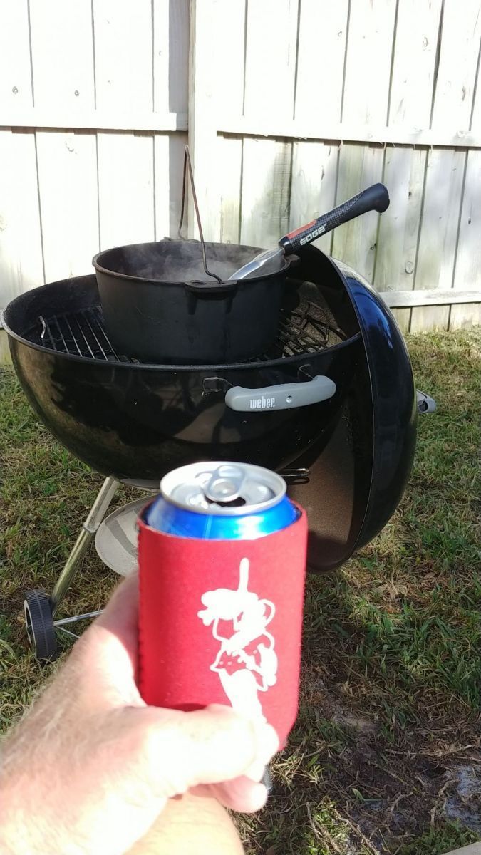Person holding a red beer can koozie with a cartoon design, next to a black charcoal grill with a dutch oven.