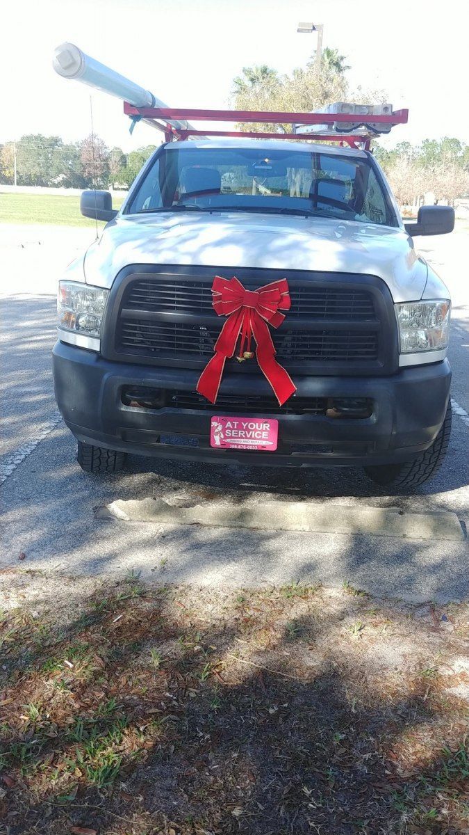White work truck with a red bow on the grill, a rack on top, and a roll of material.