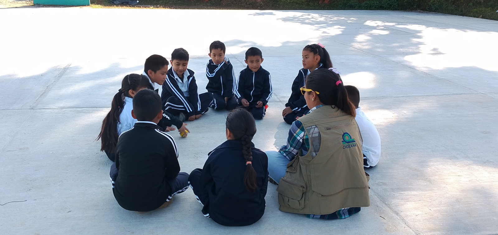 Grupo de personas sentadas en círculo al aire libre sobre una superficie iluminada por el sol, conversando entre sí.