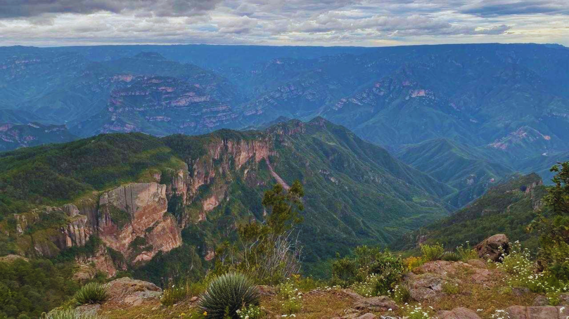 Valle de montaña con crestas verdes, acantilados rocosos y un cielo azul nublado.