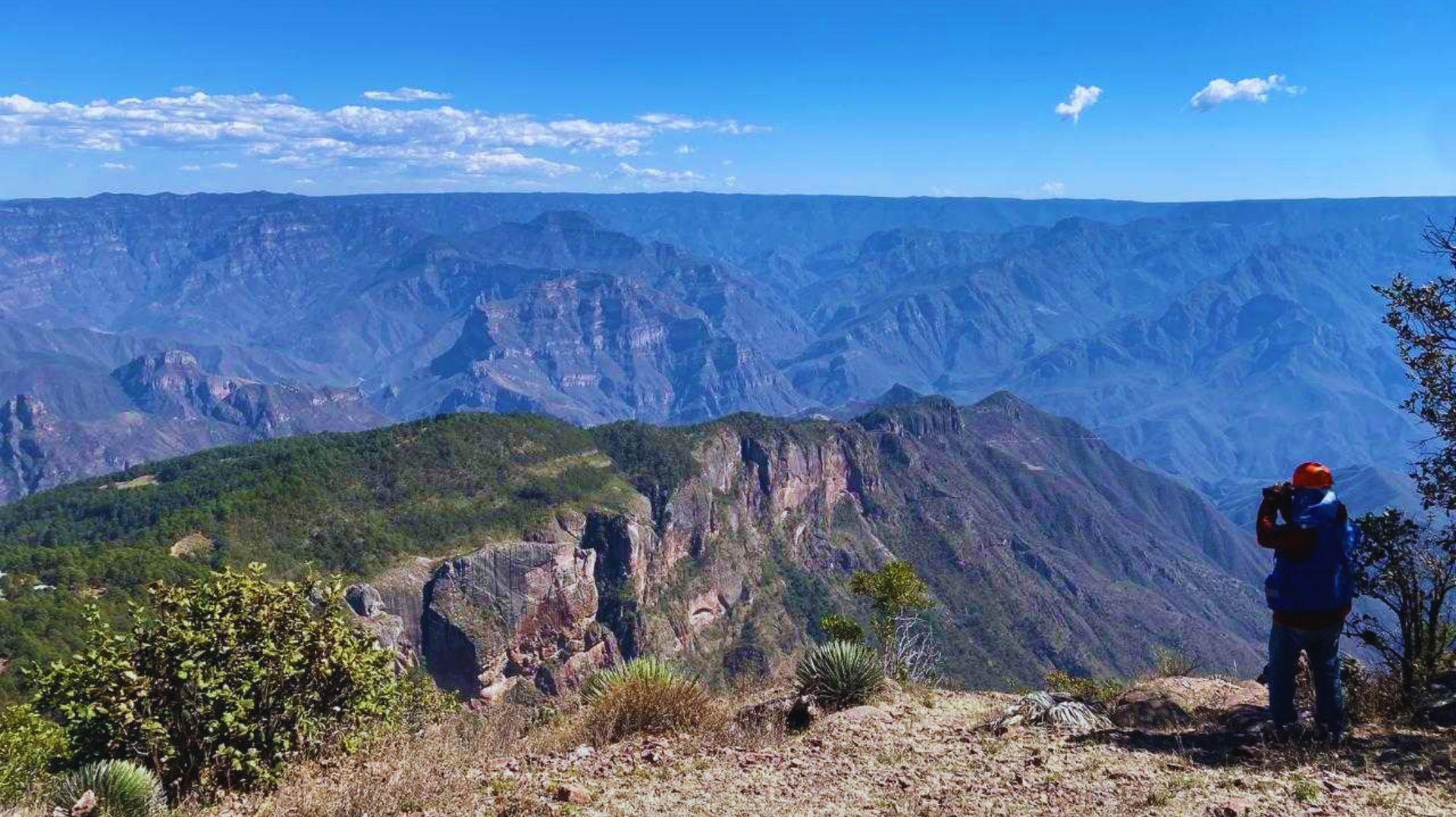 Persona con mochila contemplando un vasto cañón de montaña azul desde un mirador en lo alto de un acantilado.
