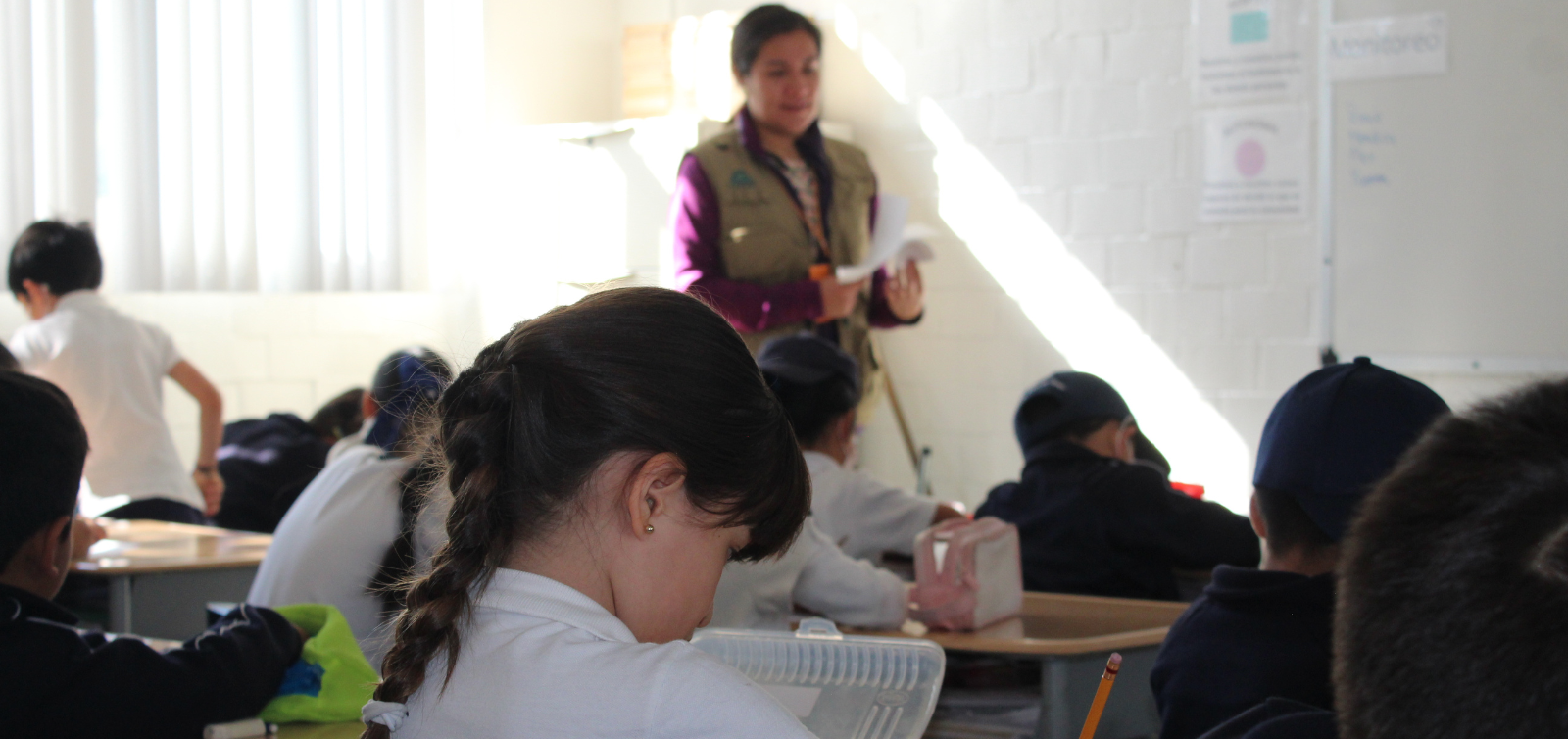 Escena de un aula con alumnos sentados en sus pupitres y un profesor de pie al frente, bajo unas ventanas luminosas.