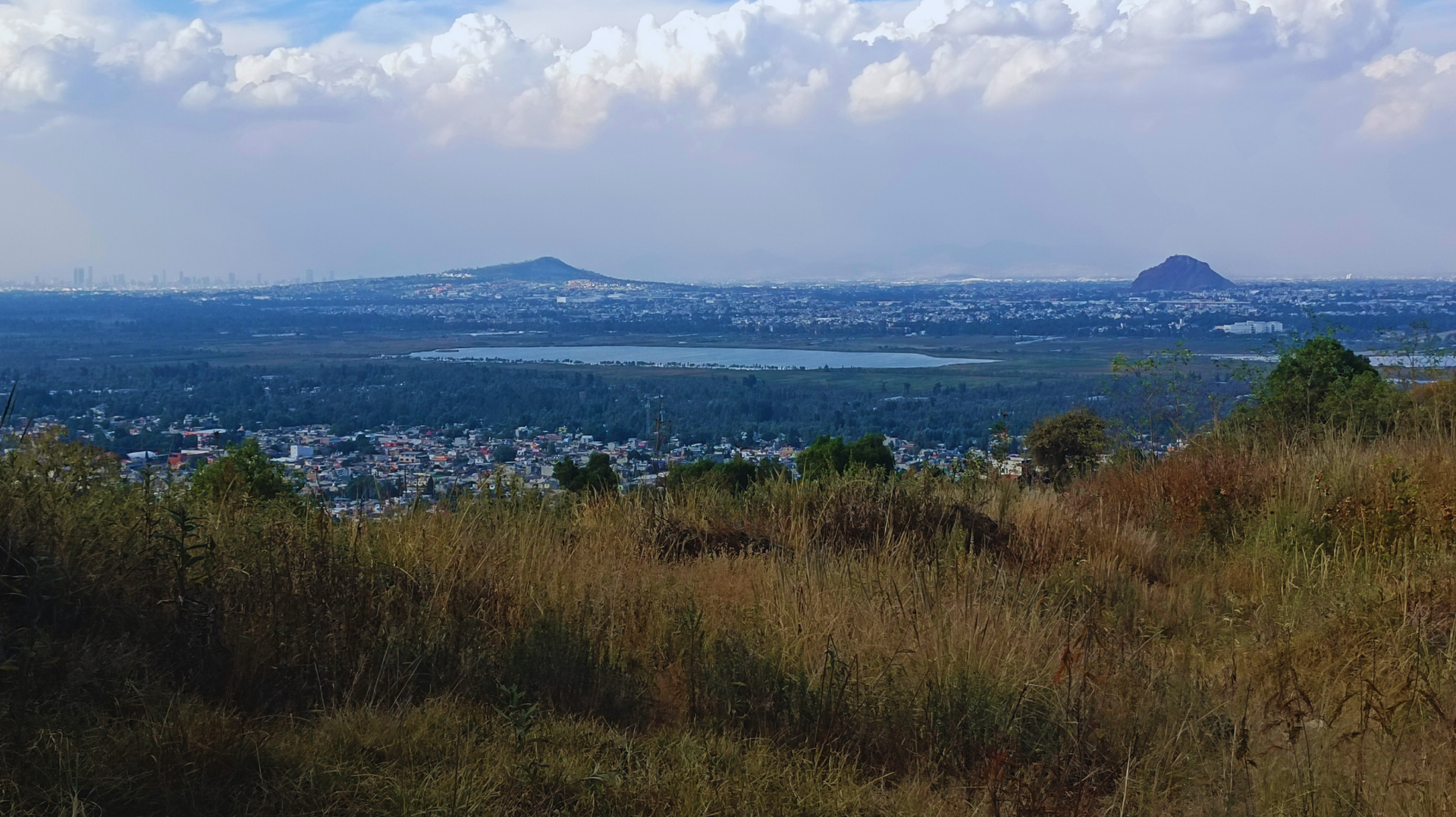 Amplia vista panorámica de un primer plano cubierto de hierba, un pueblo a lo lejos y agua azul bajo un cielo nublado.