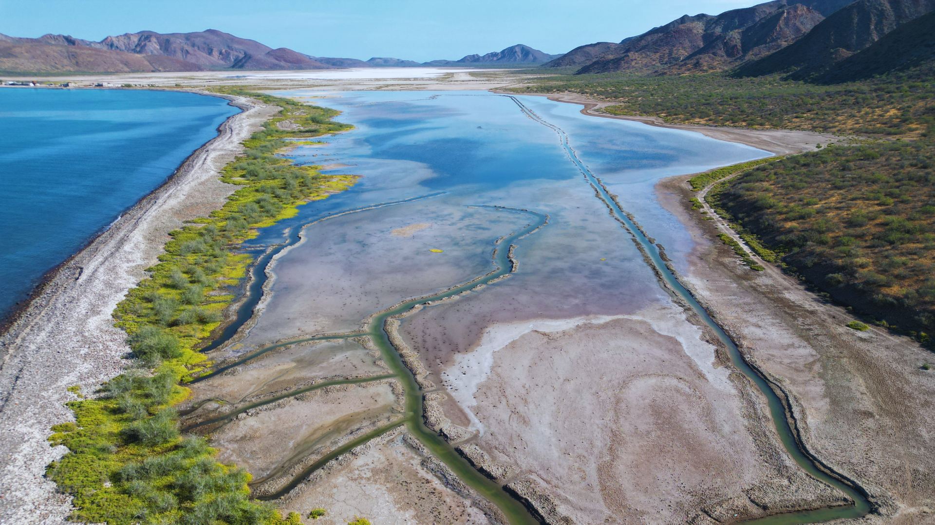Vista aérea de un humedal costero con agua azul, bancos de arena y montañas bajo un cielo despejado.