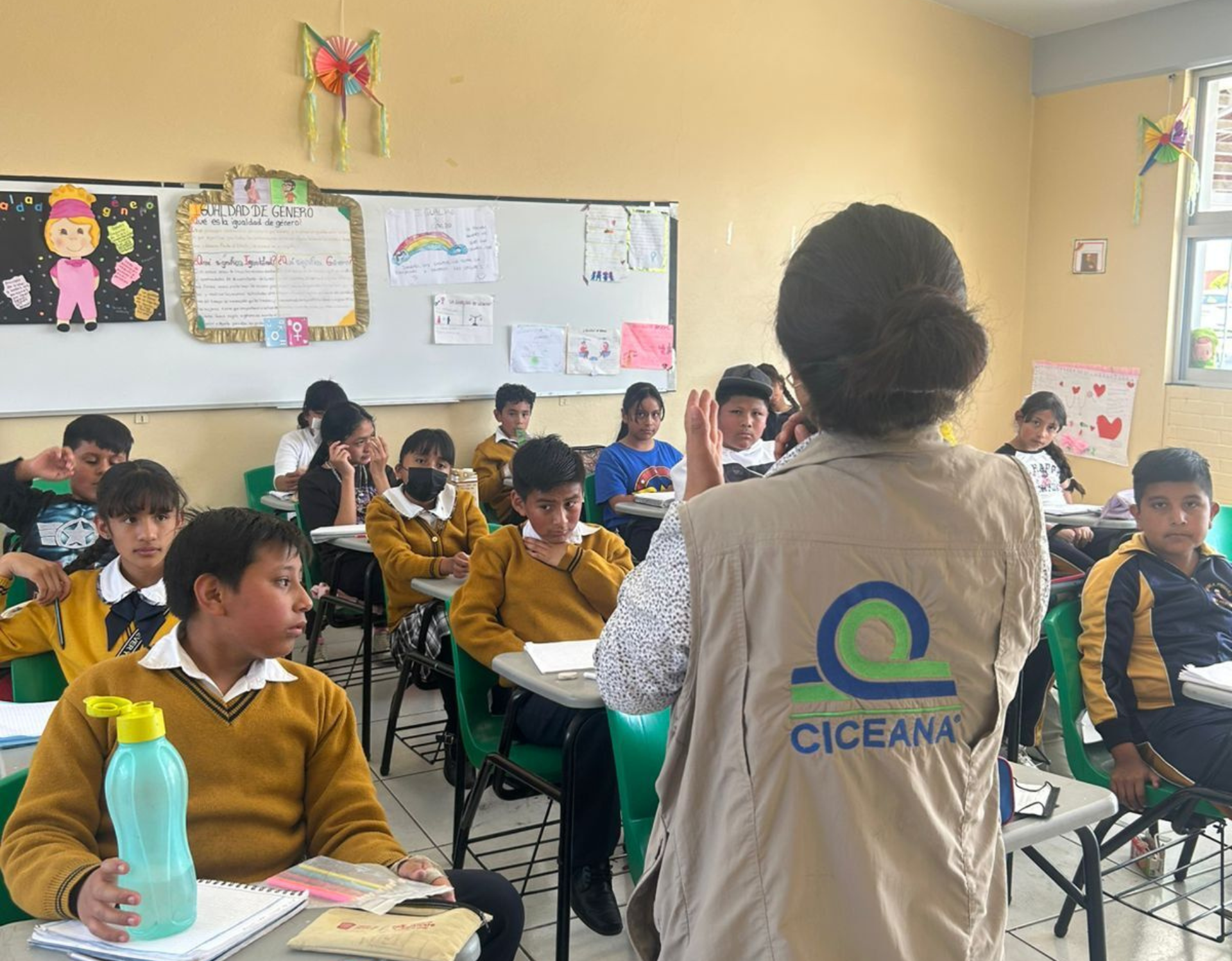 Profesora dirigiéndose a los alumnos en un aula, con estudiantes vestidos con uniformes amarillos sentados en sus pupitres.