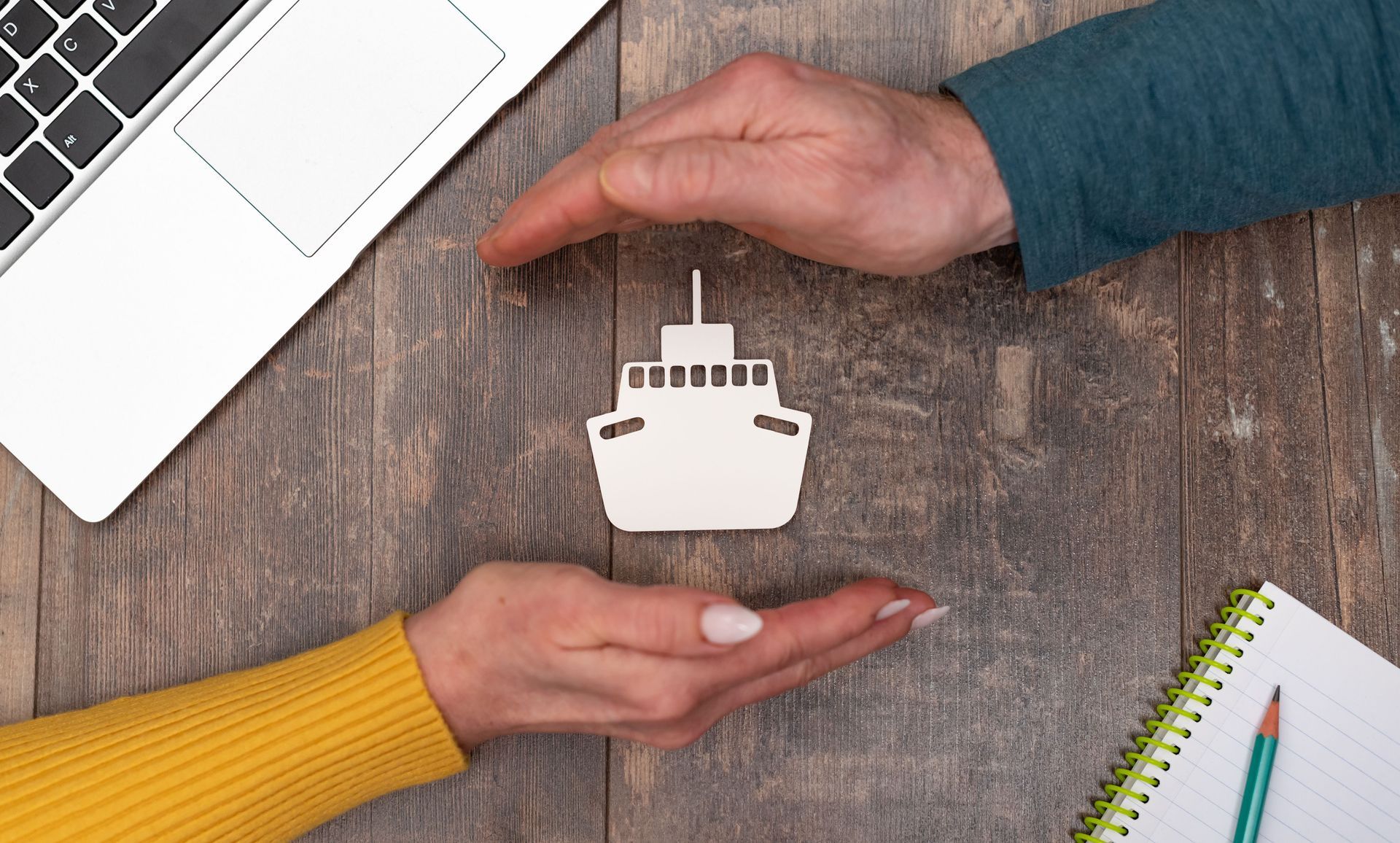 Two hands framing a small ship-shaped cutout on a desk beside a laptop and notebook. 