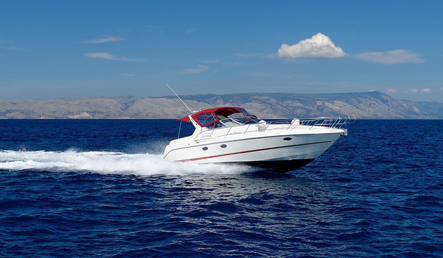 White motorboat with a red canopy cruising across open blue water past a mountain shoreline.