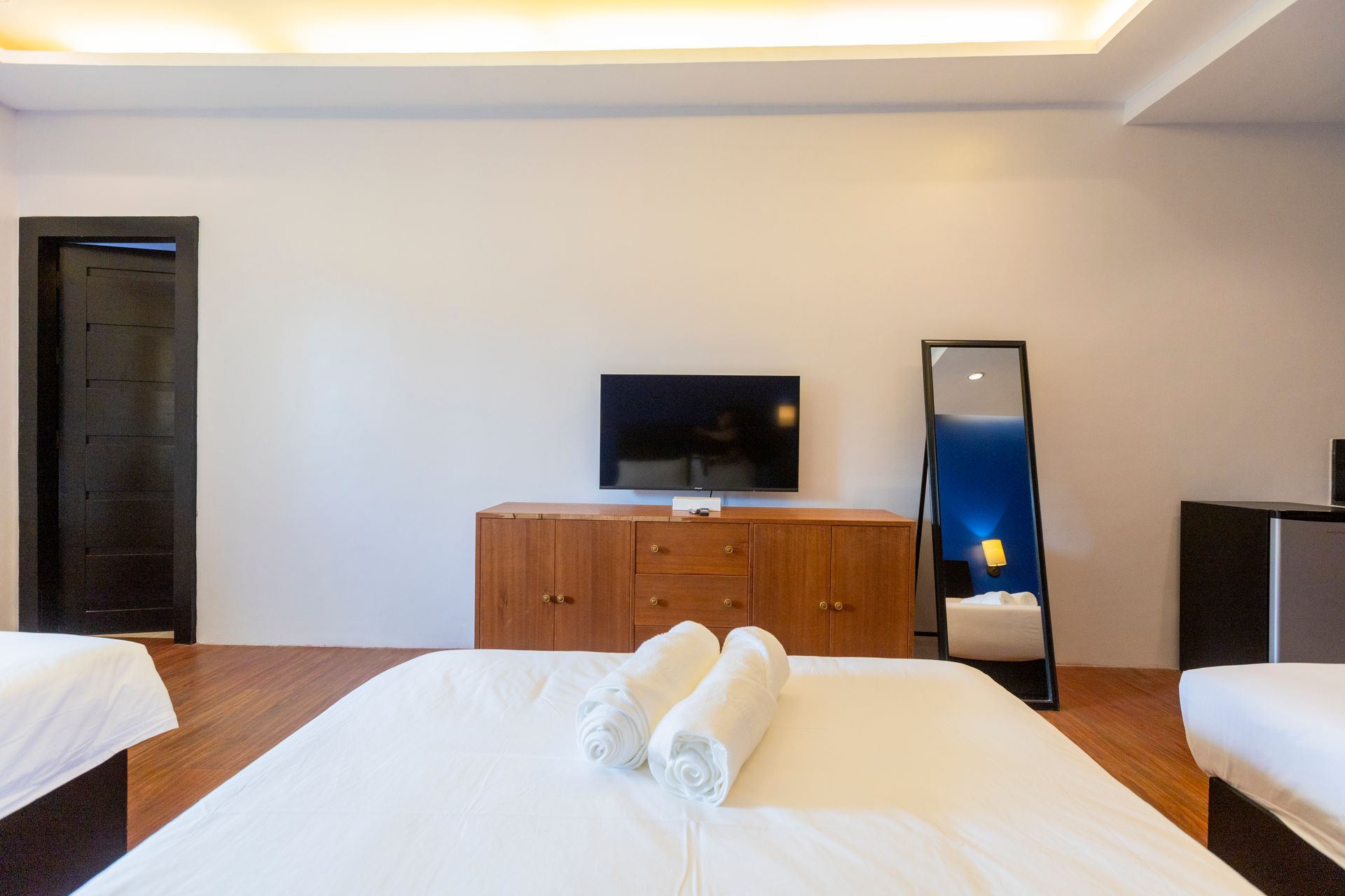 Bedroom with three beds, TV, dresser, full-length mirror, and dark door. Wooden floor.