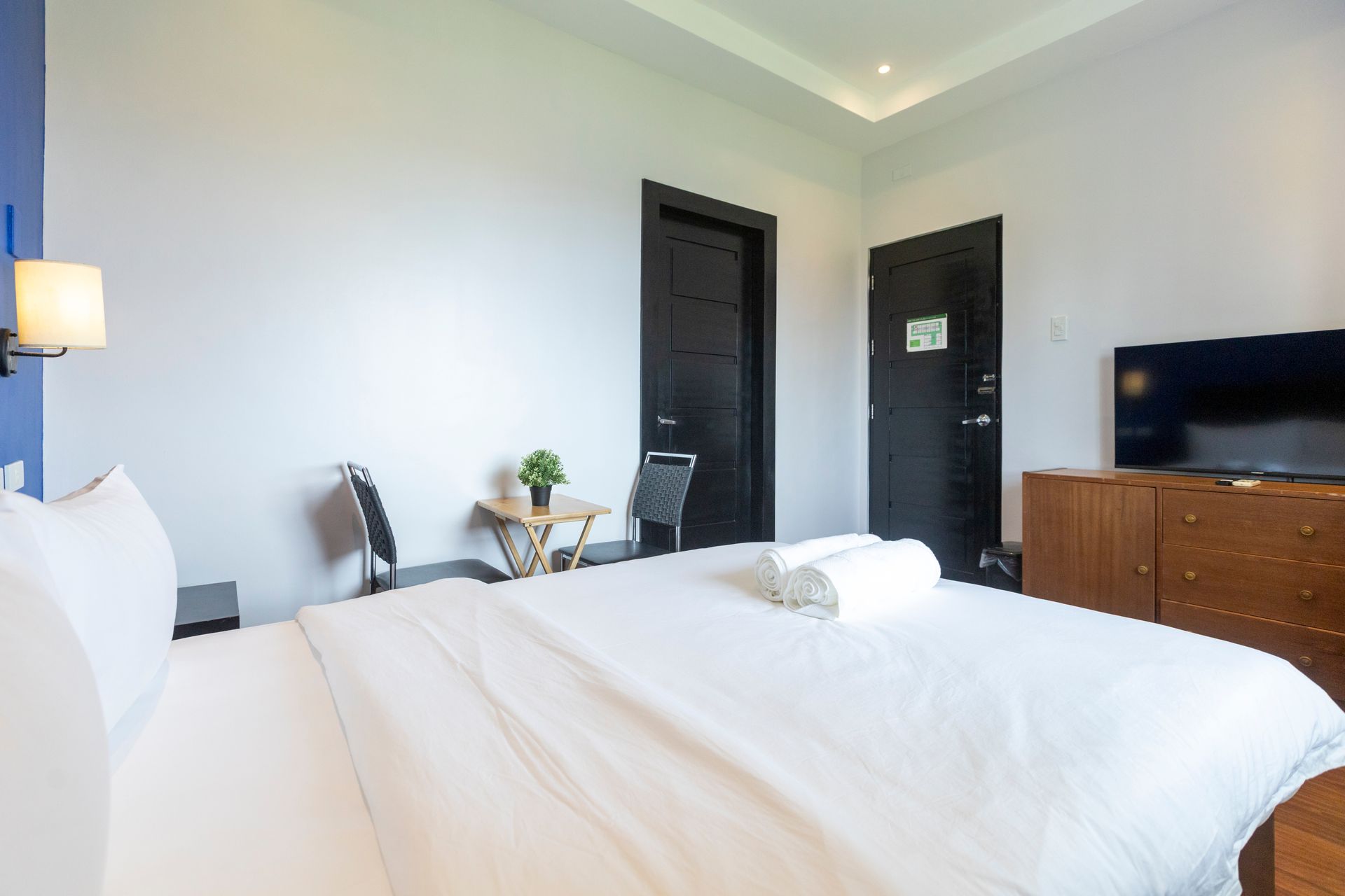 Bedroom with a bed, two chairs, small table, TV, and black doors. Bright and airy interior.