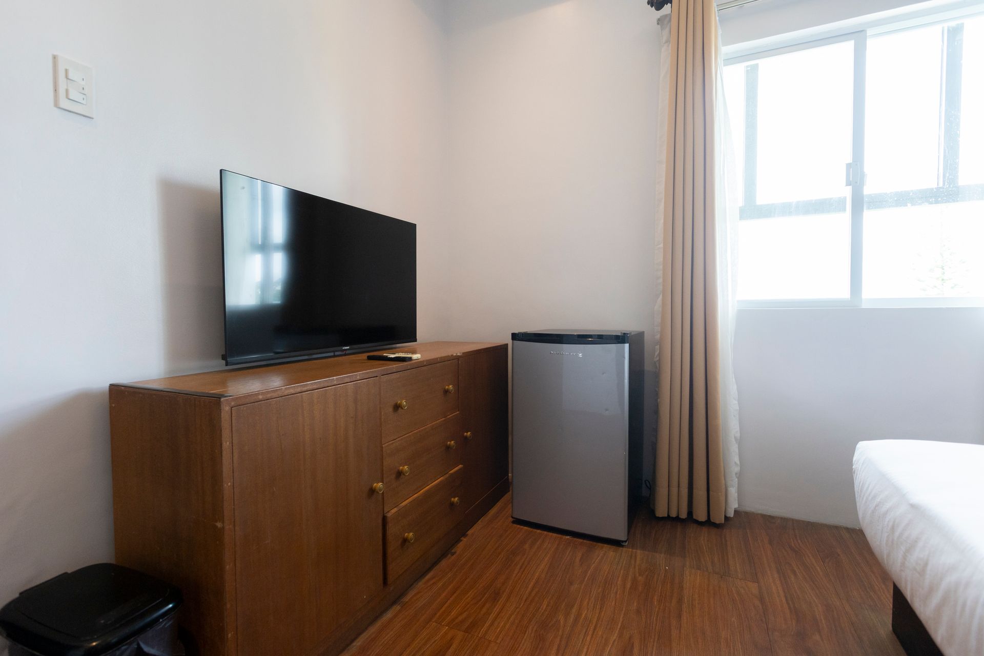 A hotel room with a TV on a wooden cabinet, a mini-fridge, and a window with curtains.