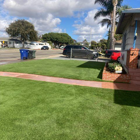 Suburban front yard with green lawn, palm trees, parked cars, and a brick house under a partly cloudy sky