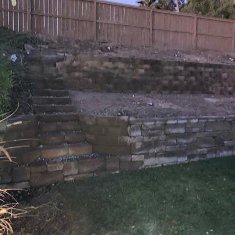 Stone retaining wall with steps leading up to a fenced backyard hillside.