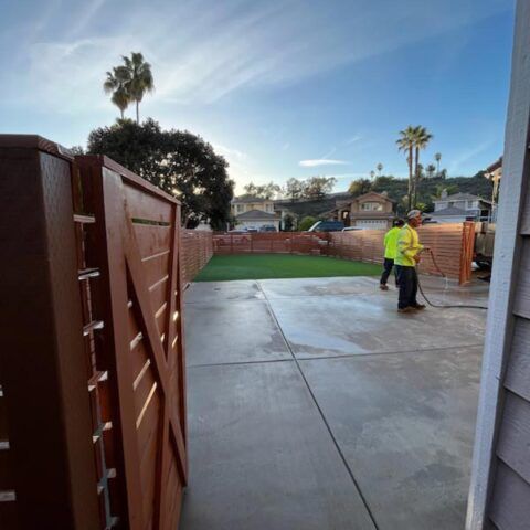 Workers in yellow vests standing on a wet concrete patio beside a fenced backyard at sunset powerwashing