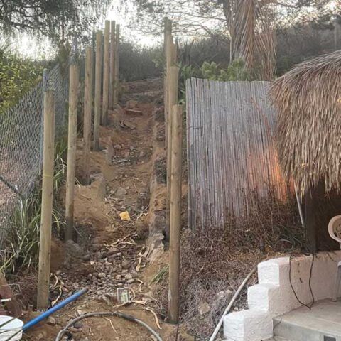 Narrow dirt stairway lined with wooden posts and corrugated fencing beside a thatched structure.