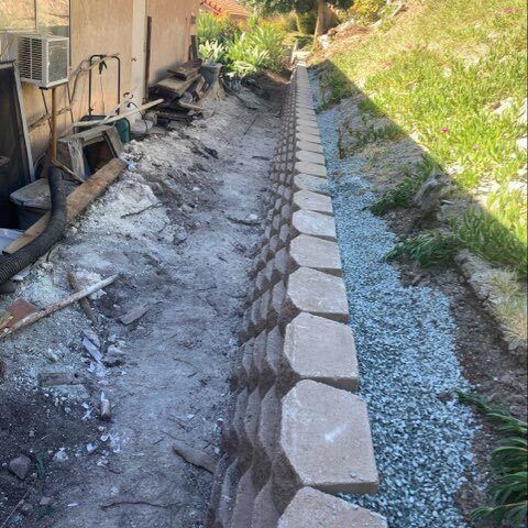 Dry retaining wall blocks line a sloped dirt trench beside a house, with gravel on the right.