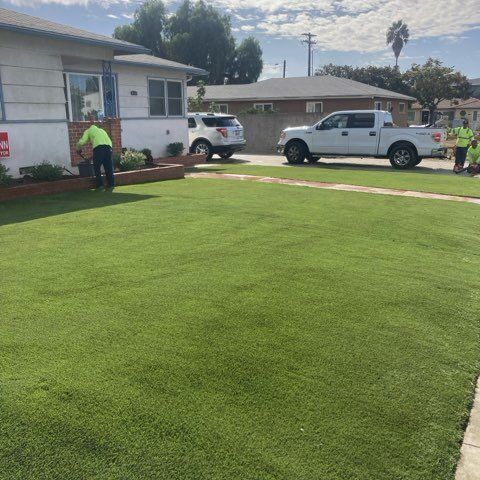 Workers mowing a bright green lawn beside a white house with trucks parked in the driveway.