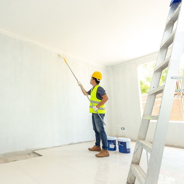 Painter in a hard hat and safety vest rolls white paint on a wall in a bright room beside a ladder.
