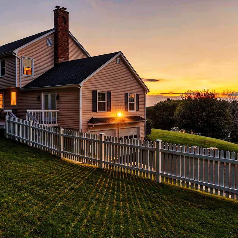Sunset over a coastal house with a white fence and lit windows on a grassy slope
