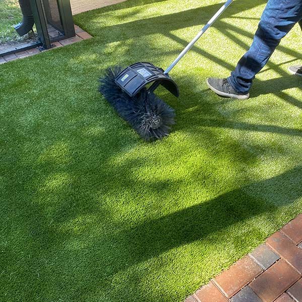 Person using a push spreader on a green lawn near a brick path and fence