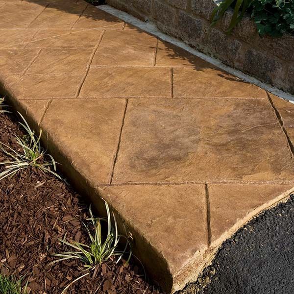 Tan stamped concrete corner beside dark asphalt and mulch with small plants