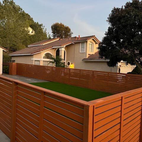 Backyard with modern wooden fence, green artificial turf, and a beige house at sunset