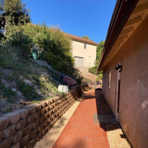 Narrow red-tiled outdoor walkway beside a stucco building and hillside retaining wall under a blue sky