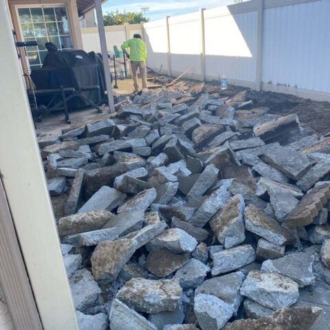 Backyard with a large pile of broken concrete stones and a worker near a fence.