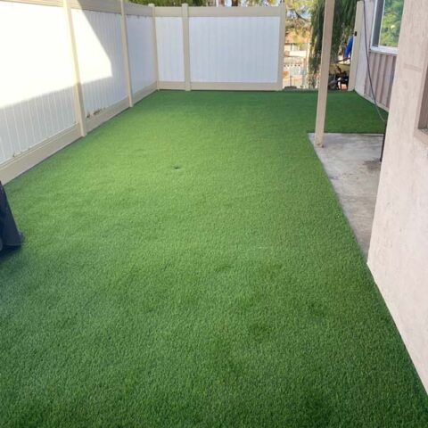Narrow fenced side yard with bright green artificial turf and a concrete walkway along the right side.