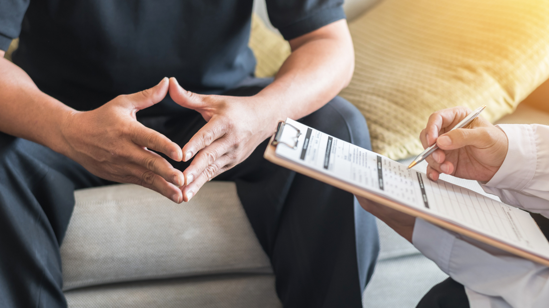 A man is sitting on a couch talking to a doctor who is holding a clipboard.