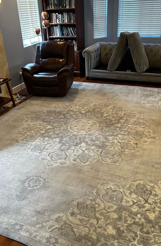 A living room with a large, patterned light-gray rug, a brown leather recliner, a gray tufted sofa, and a tall bookshelf.