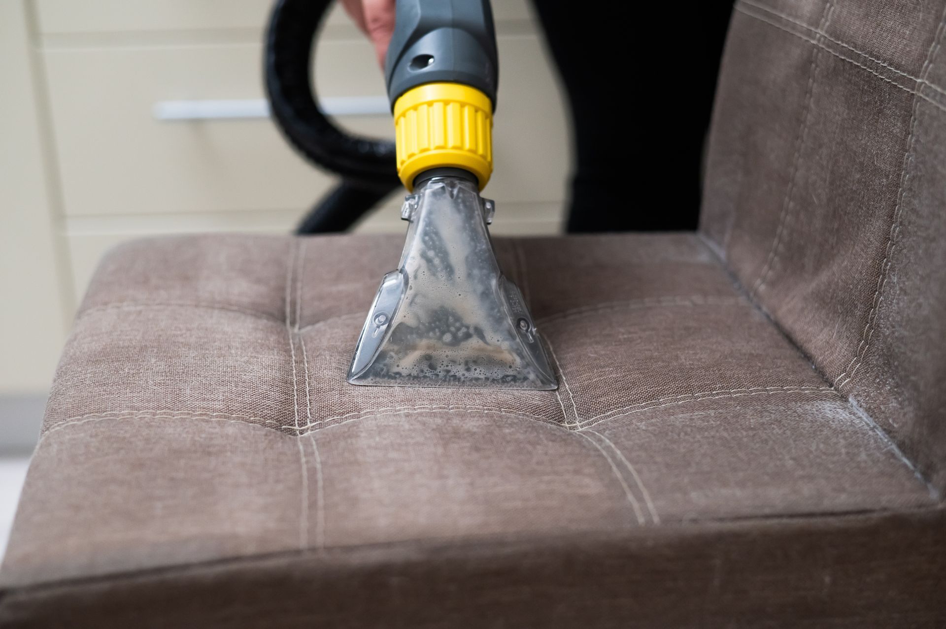 A hand uses a steam cleaning upholstery tool to clean a brown fabric chair.