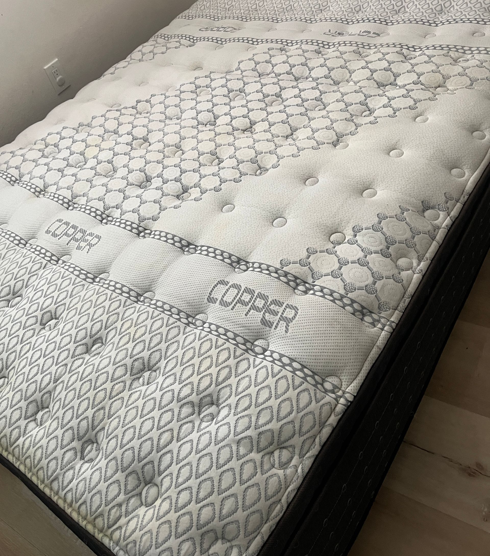 A mattress with a tufted, white and gray patterned fabric top sitting on a black bed frame in a room.