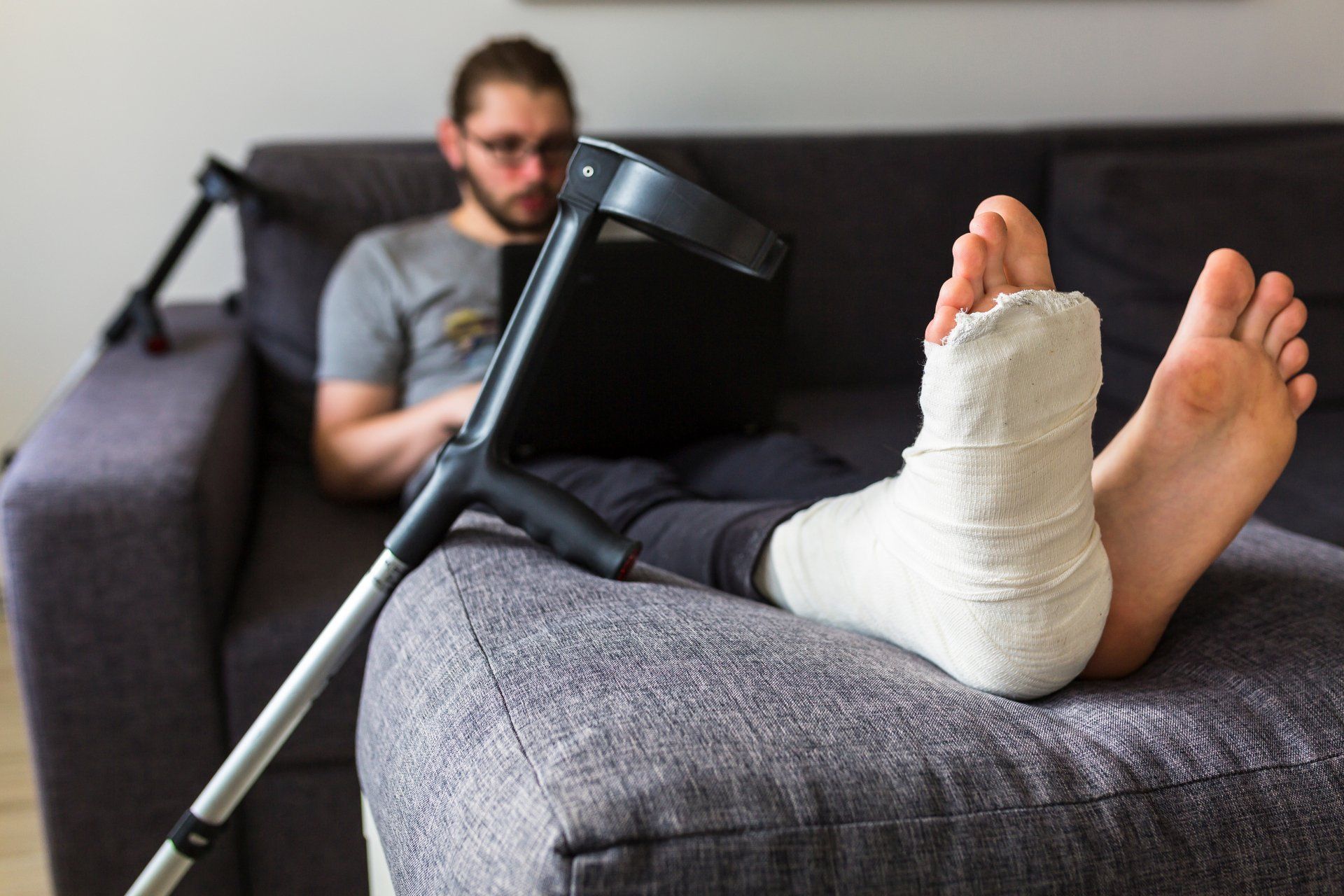 Man with a leg cast on a couch, using a laptop with crutch nearby. Man with a leg cast on a couch, using a laptop with crutch nearby.
