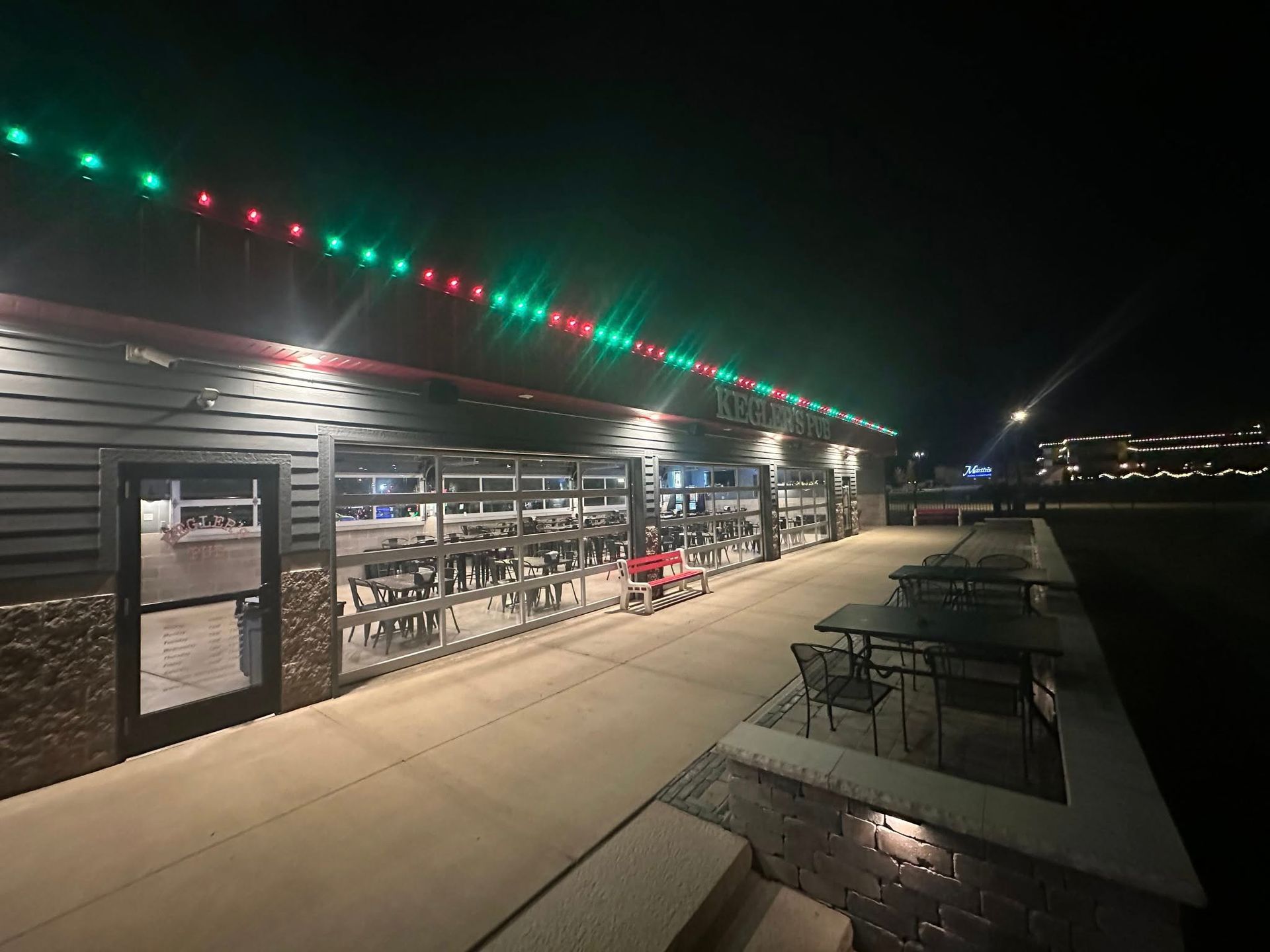 Outdoor restaurant patio at night with red and green string lights and empty tables along a walkway