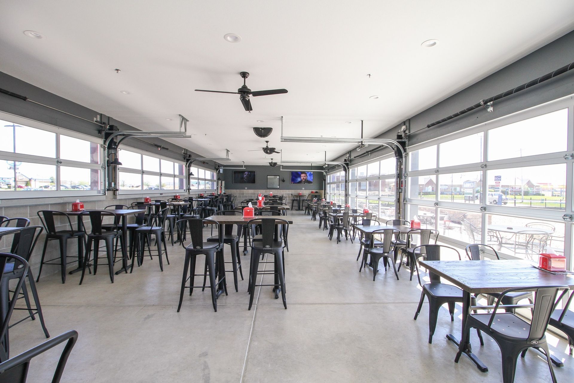 Bright, empty restaurant dining area with black tables and stools, large windows, and a TV on the back wall