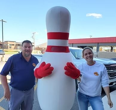Two people posing with a giant bowling pin mascot outside a building
